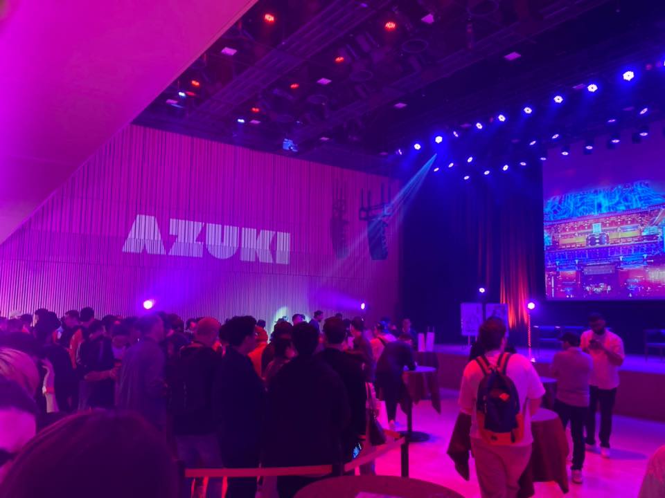 ライブイベント会場の内部。紫とピンクの照明に照らされた空間。壁に「AZUKI」という大きな文字が見える。ステージと大型スクリーンが見え、多くの観客が集まっている。