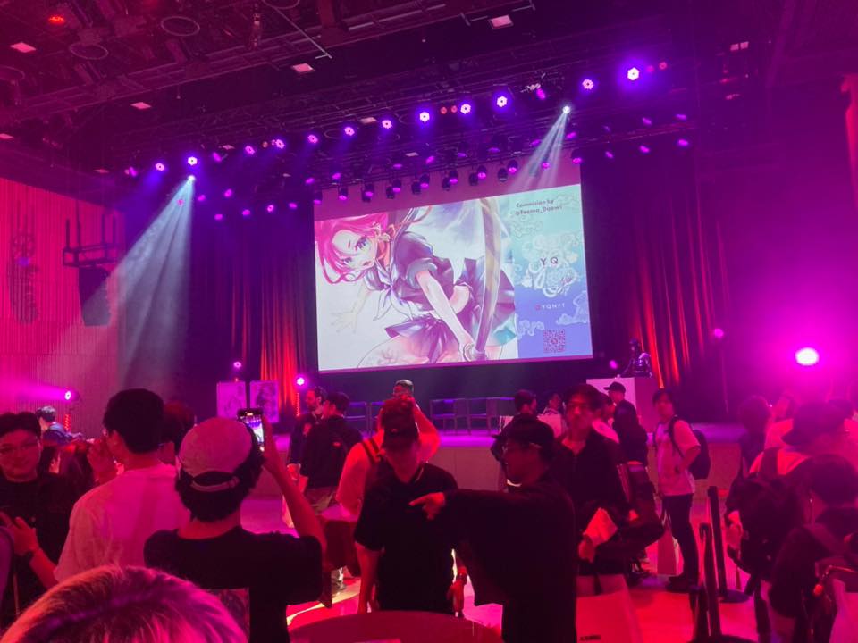 ライブイベント会場のステージ。大型スクリーンにアニメキャラクターが映し出されている。紫とピンクの照明が強く、観客が前方に集まっている。