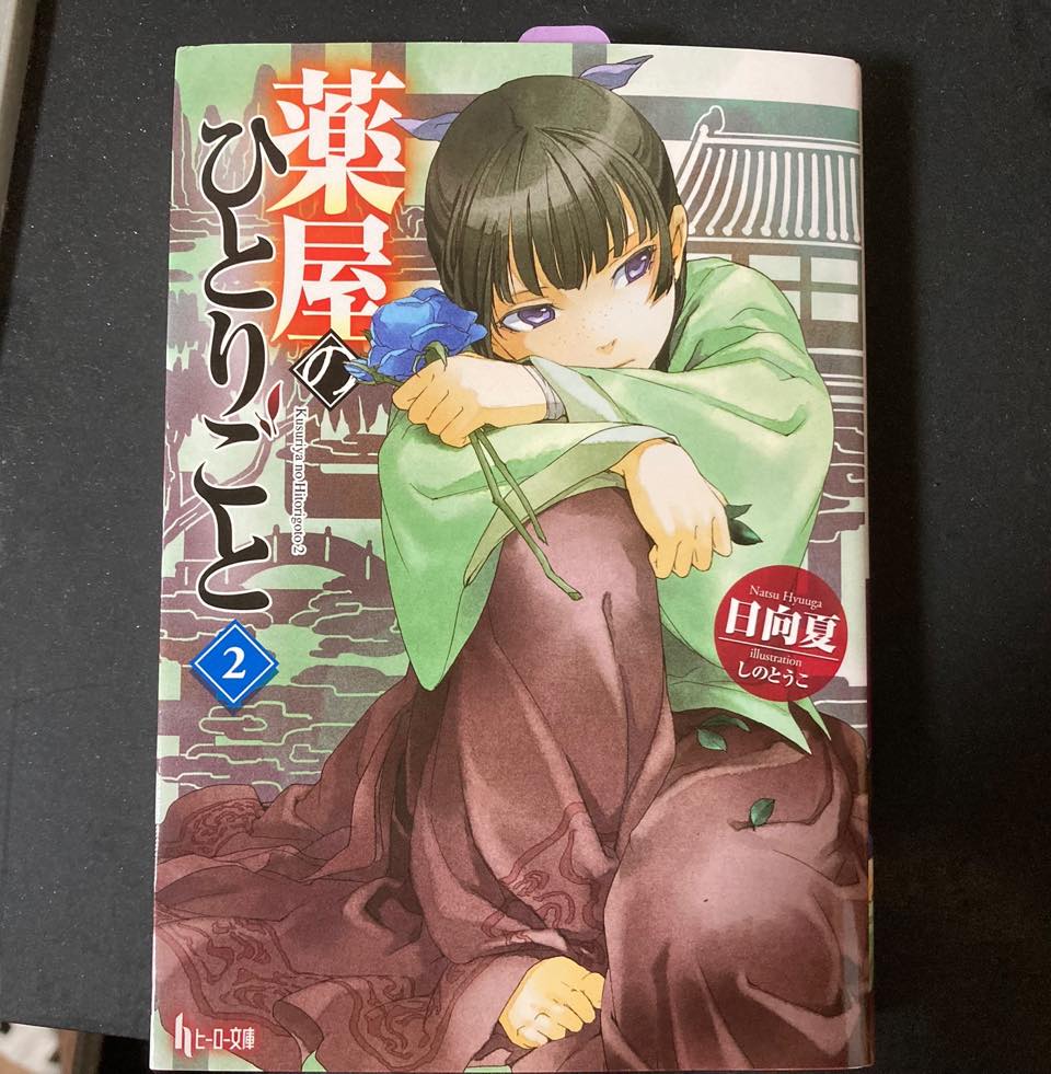 『薬屋のひとりごと』第2巻の表紙。緑の上着と紫色の着物を着た少女が青い花を持ち、腕に顎を乗せてじっと見つめている。背景には東洋風の建物が描かれている。タイトルは赤色で大きく表示され、作者は日向夏、イラストはしのとうこと記されている。