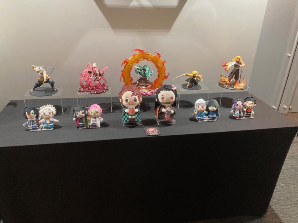 この画像はアニメやビデオゲームシリーズのフィギュアやグッズのコレクションを展示しています。アイテムは黒いディスプレイスタンドに配置されています。後列には、アクションポーズをとるキャラクターの大きくて詳細なフィギュアがあります。前列には様々なキャラクターのより小さなちびスタイルのフィギュアが並んでいます。リアルなものからカートゥーン調のものまで、スタイルが混在しています。展示は展示会や小売店のような場所にあるようです。