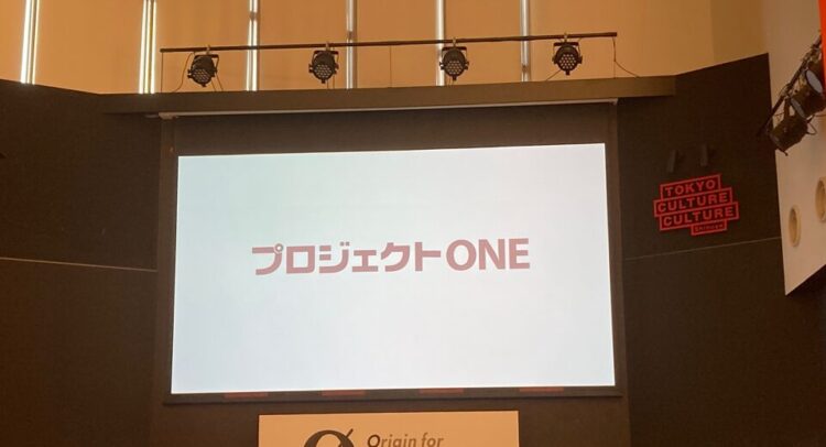 スクリーンに「プロジェクトONE」と書かれたプレゼンテーションスライドが映し出されている。スライドは白い背景に赤い文字で「プロジェクトONE」と書かれており、場所は「TOKYO CULTURE CULTURE」と書かれたサインがある渋谷のイベントスペース。