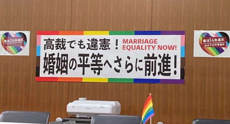 この画像は、結婚の平等を求めるメッセージが掲げられた会場の様子を示しています。大きな横断幕には「高裁でも違憲！結婚の平等へさらに前進！ Marriage Equality Now!」と書かれており、LGBTQ+コミュニティの結婚平等を求める訴えを示す内容です。背景には憲法第24条や第14条への言及があり、法の下の平等と結婚の平等を主張しています。また、虹色のフラッグが会場内に置かれており、LGBTQ+のシンボルを用いて結婚の平等に対する支援を表しています。