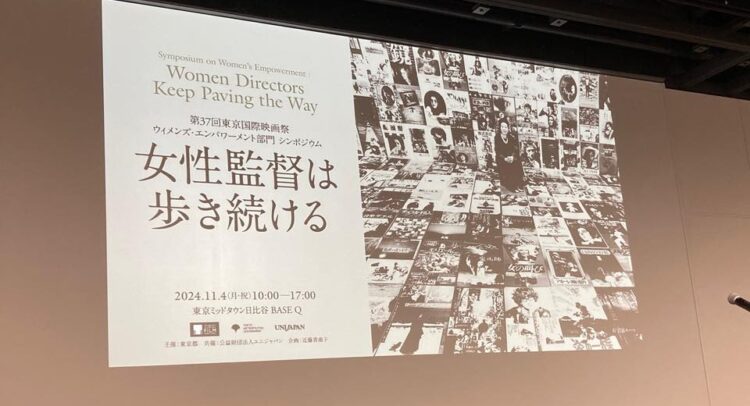 この画像は、第37回東京国際映画祭におけるシンポジウム「女性監督は歩き続ける」に関するポスターまたはスクリーン投影のようです。テーマは「Women Directors Keep Paving the Way」で、ウィメンズ・エンパワメント部門のイベントとして開催されています。日付は2024年11月4日（月・祝）、場所は東京ミッドタウン日比谷のBASE Qで、開催時間は10:00〜17:00となっています。背景には映画関連のポスターが敷き詰められ、女性の姿が写っているモノクロの写真が使われています。