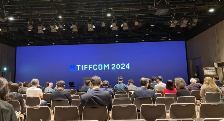 TIFFCOM 2024の会場内部の様子を撮影した写真。観客席からスクリーンを正面に捉えた構図で、スクリーンには「TIFFCOM 2024」と白文字で表示されています。座席には多くの参加者が着席しており、会場全体は落ち着いた照明が施され、プロフェッショナルな雰囲気を醸し出しています。上部にはスポットライトや音響設備が確認できます。