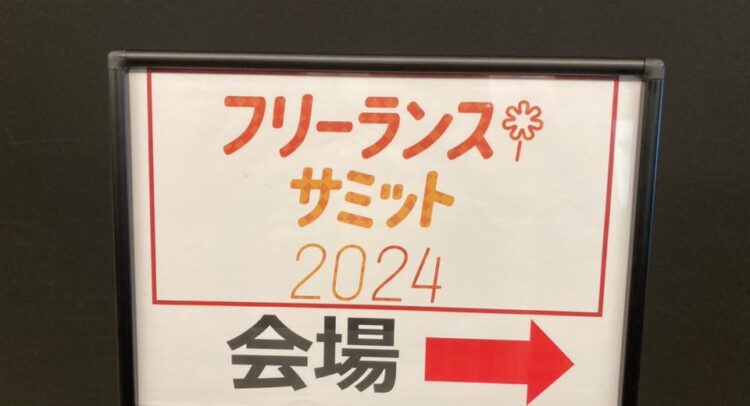 「フリーランス サミット 2024 会場」と書かれた案内看板。看板は白地に赤とオレンジ色の文字で構成され、右方向を示す大きな赤い矢印が目立つデザイン。黒い枠に収められ、背景は暗めの壁。