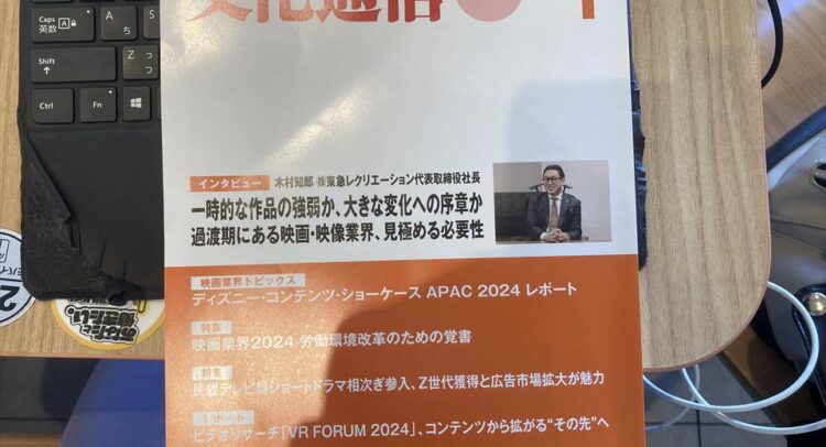 「文化通信ジャーナル 2025年1月号」の表紙。赤と白を基調としたデザインで、「映画・放送・音楽の業界誌」というキャッチコピーが記載されている。特集インタビューでは、俳優の木村規朗氏のインタビューが掲載されており、「一時的な作品の強弱か、大きな変化への序章か　過渡期にある映画・映像業界、見極める必要性」というタイトルが目立つ。左にはパソコンのキーボード、右には木目調のテーブルとバッグが映り込んでいる。